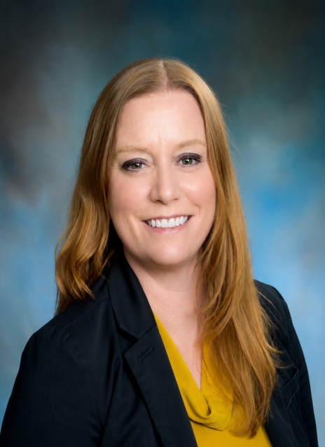 Headshot of a smiling woman in a dark blazer and yellow top against a blue studio backdrop