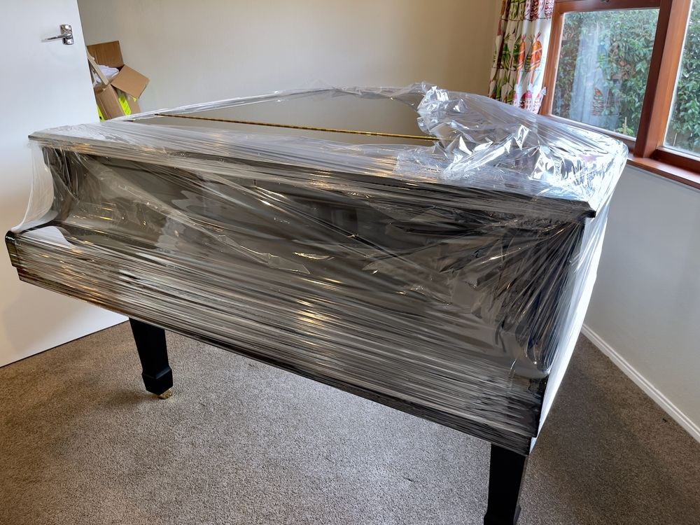 Wrapped black grand piano in a room near windows, covered in plastic for moving or storage