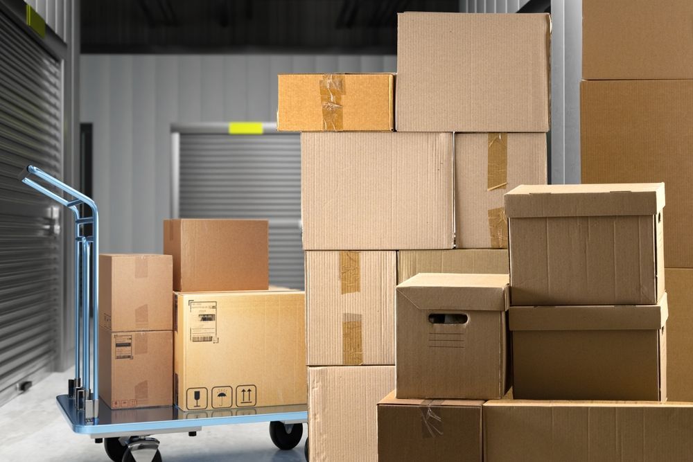 Stacked cardboard boxes in a warehouse, with a blue hand truck and metal storage shelves.