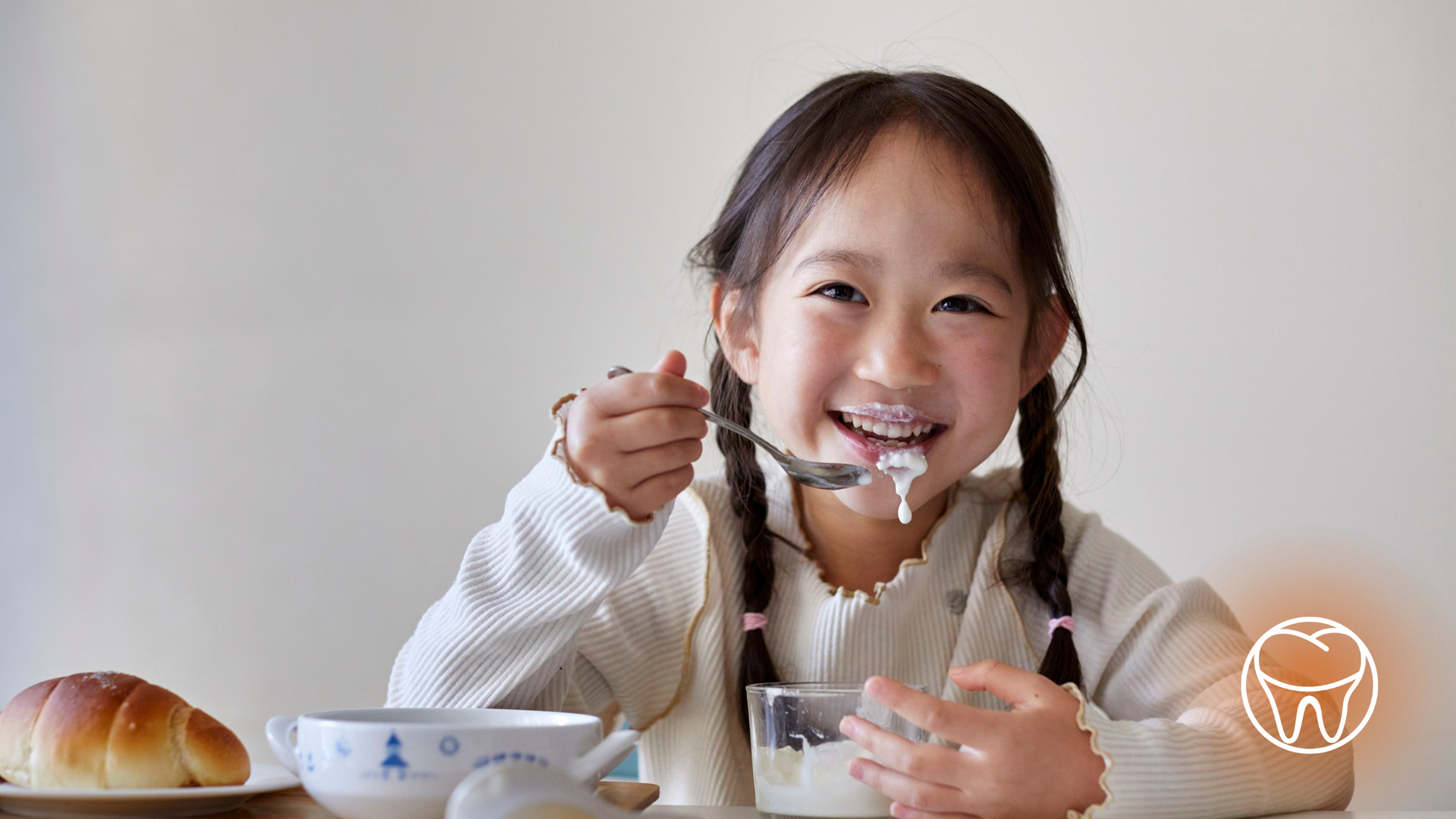 Girl smiling while eating yogurt with a croissant and cup on the table.