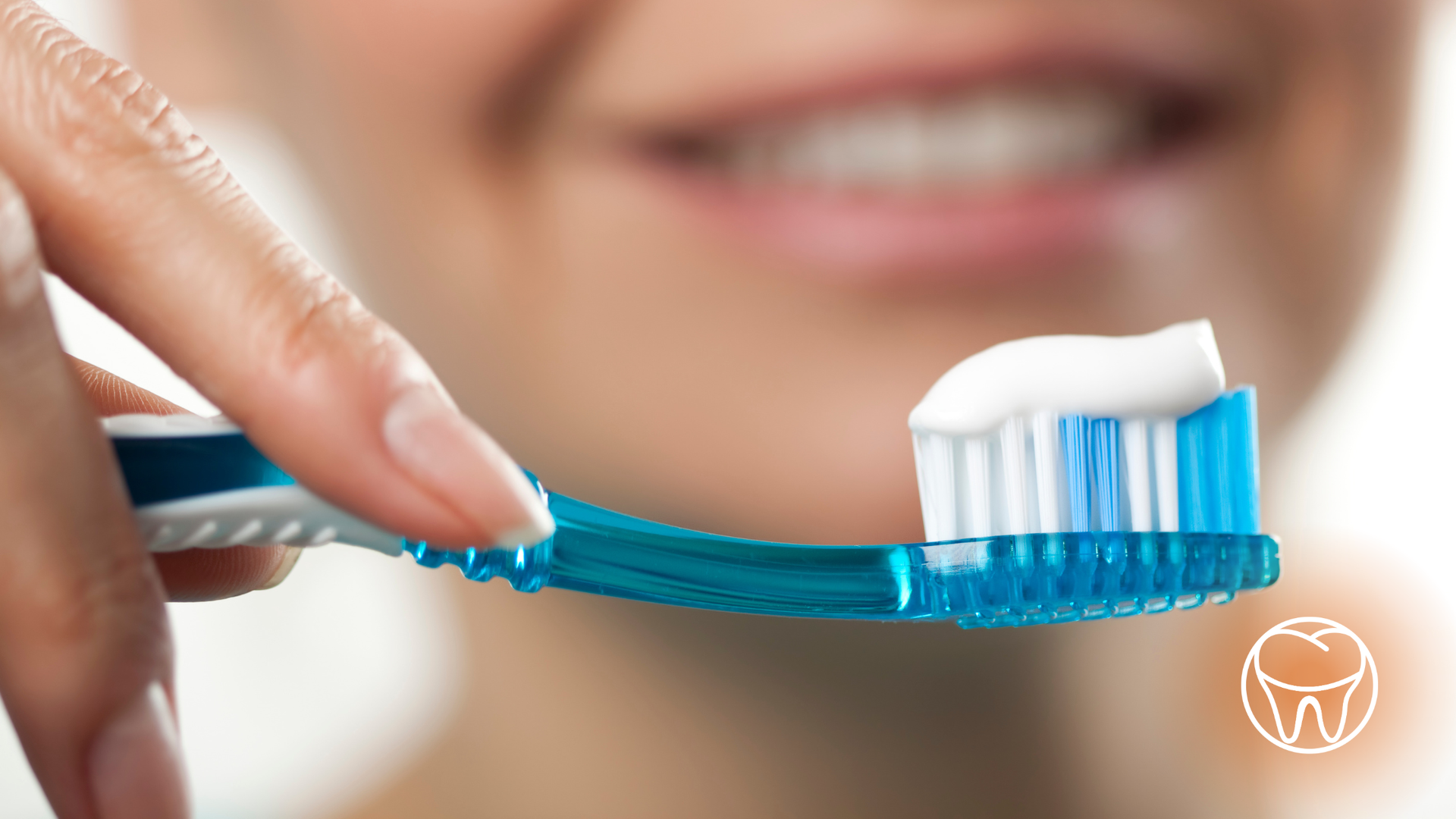 A hand holding a blue toothbrush with toothpaste, smiling person in background.