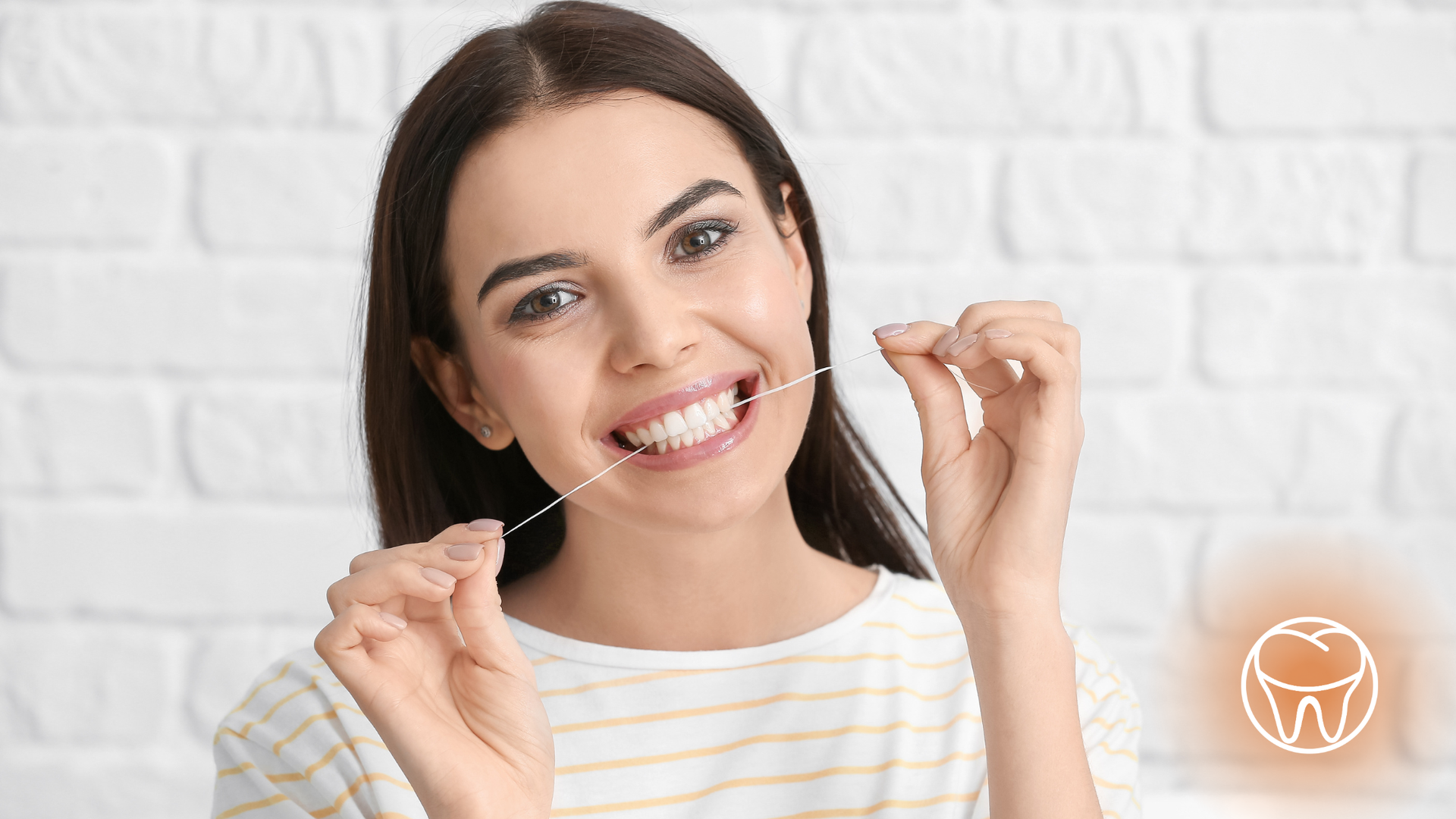 Woman flossing teeth, smiling at camera against a white brick wall.