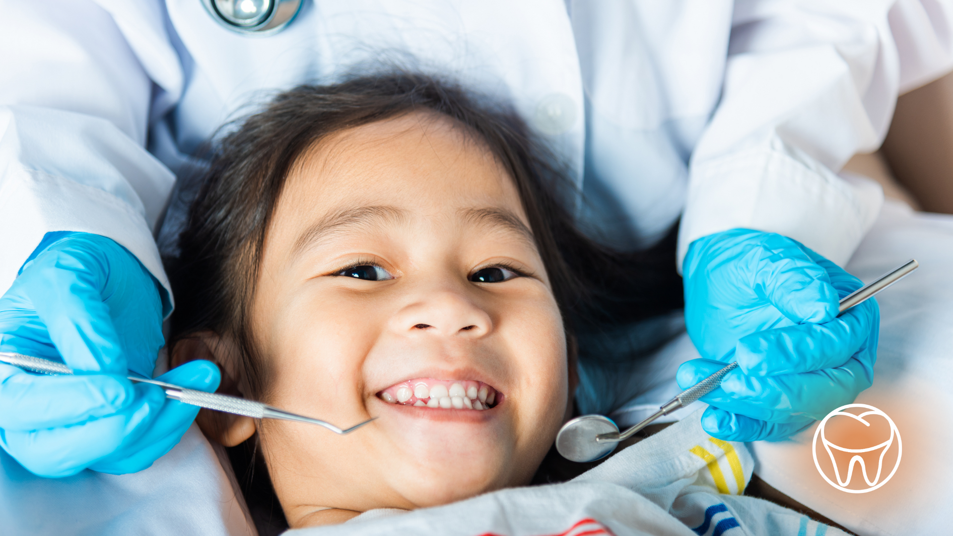 Child smiling at dentist with tools, dentist wearing gloves and a lab coat.