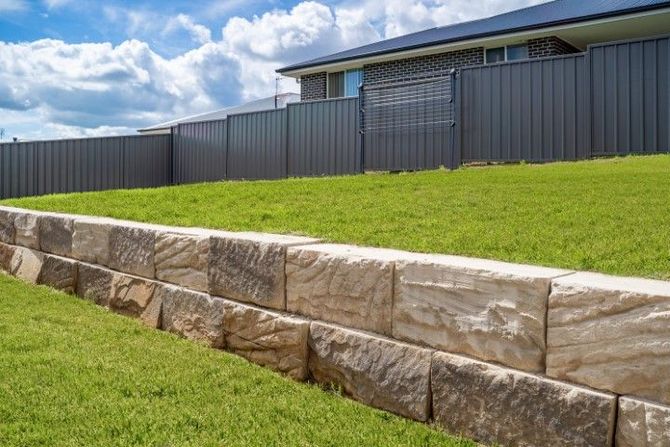 Stone retaining wall beside a green lawn and dark fence under a blue sky with clouds