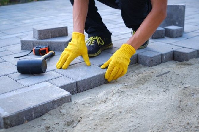Worker in yellow gloves laying gray paving stones on a patio.