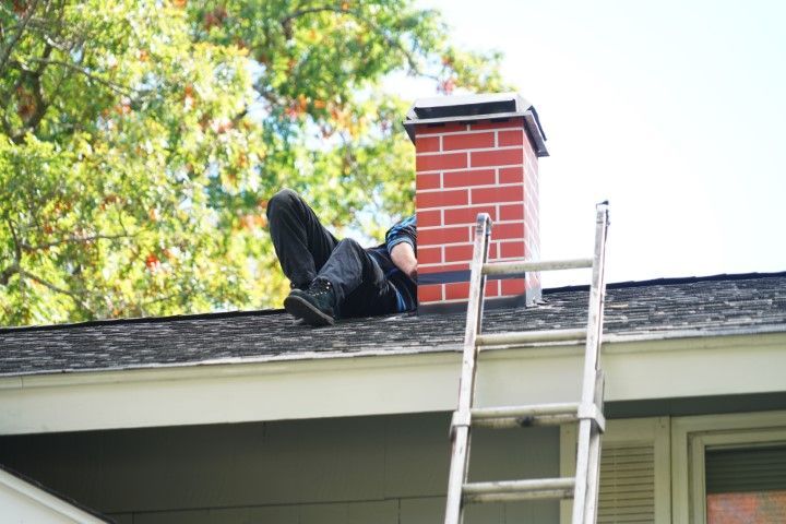 Worker on a roof beside a red brick chimney, with a ladder leaning up from the eaves.