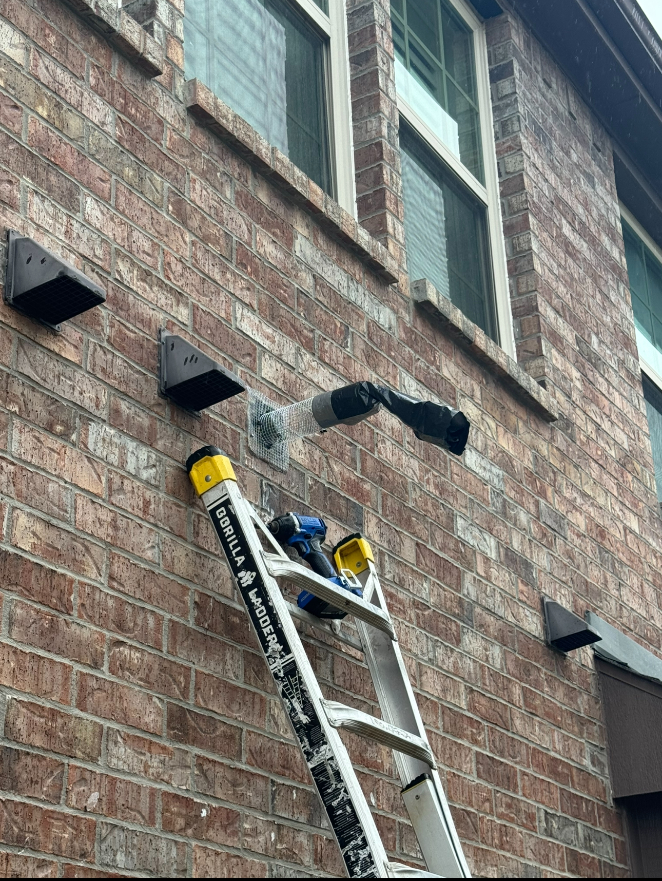 A person on a ladder drills into a brick wall next to two brackets below a window.