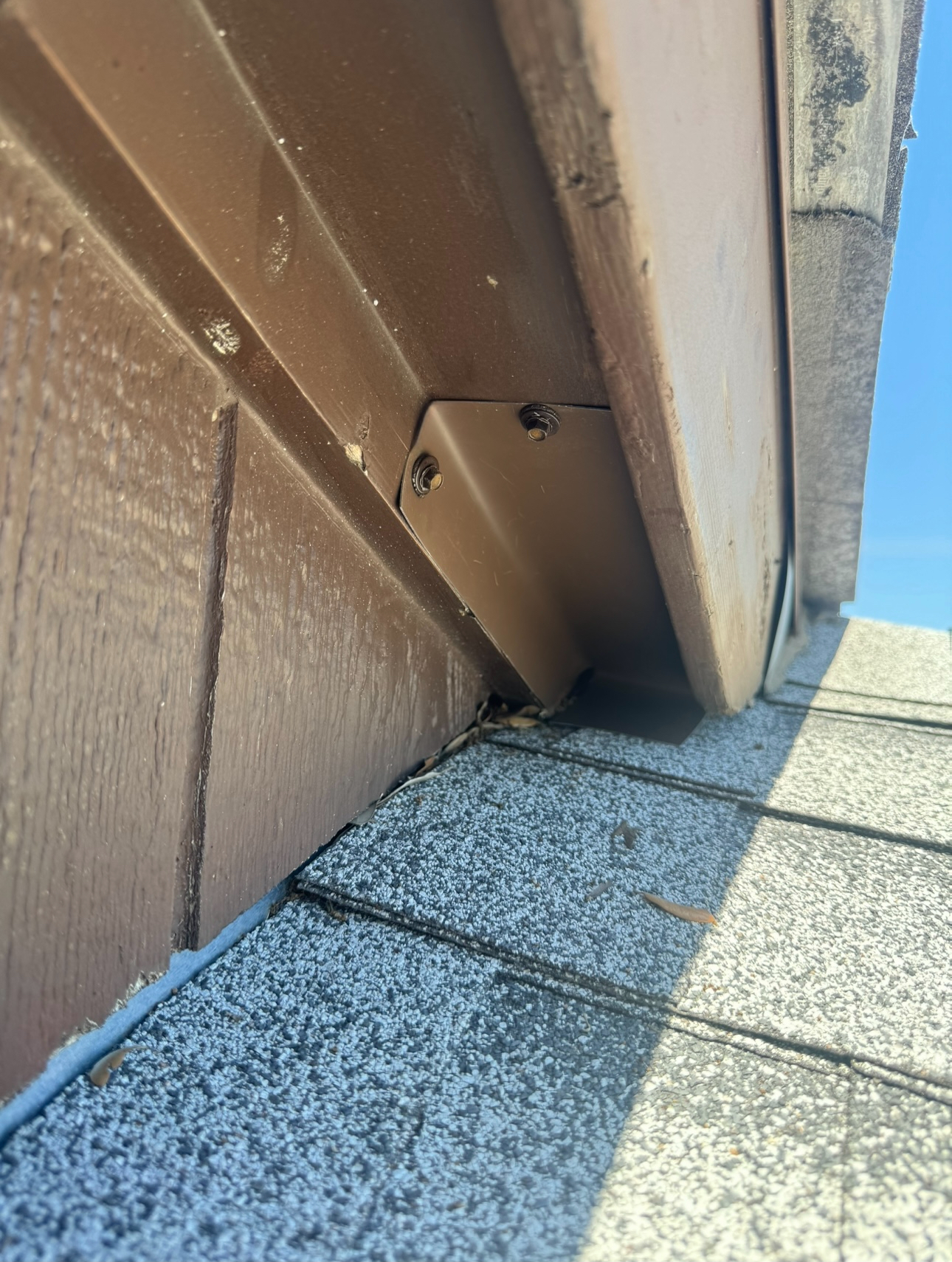 Brown siding and soffit meet asphalt shingles. A brown metal bracket is attached. Sunny day.