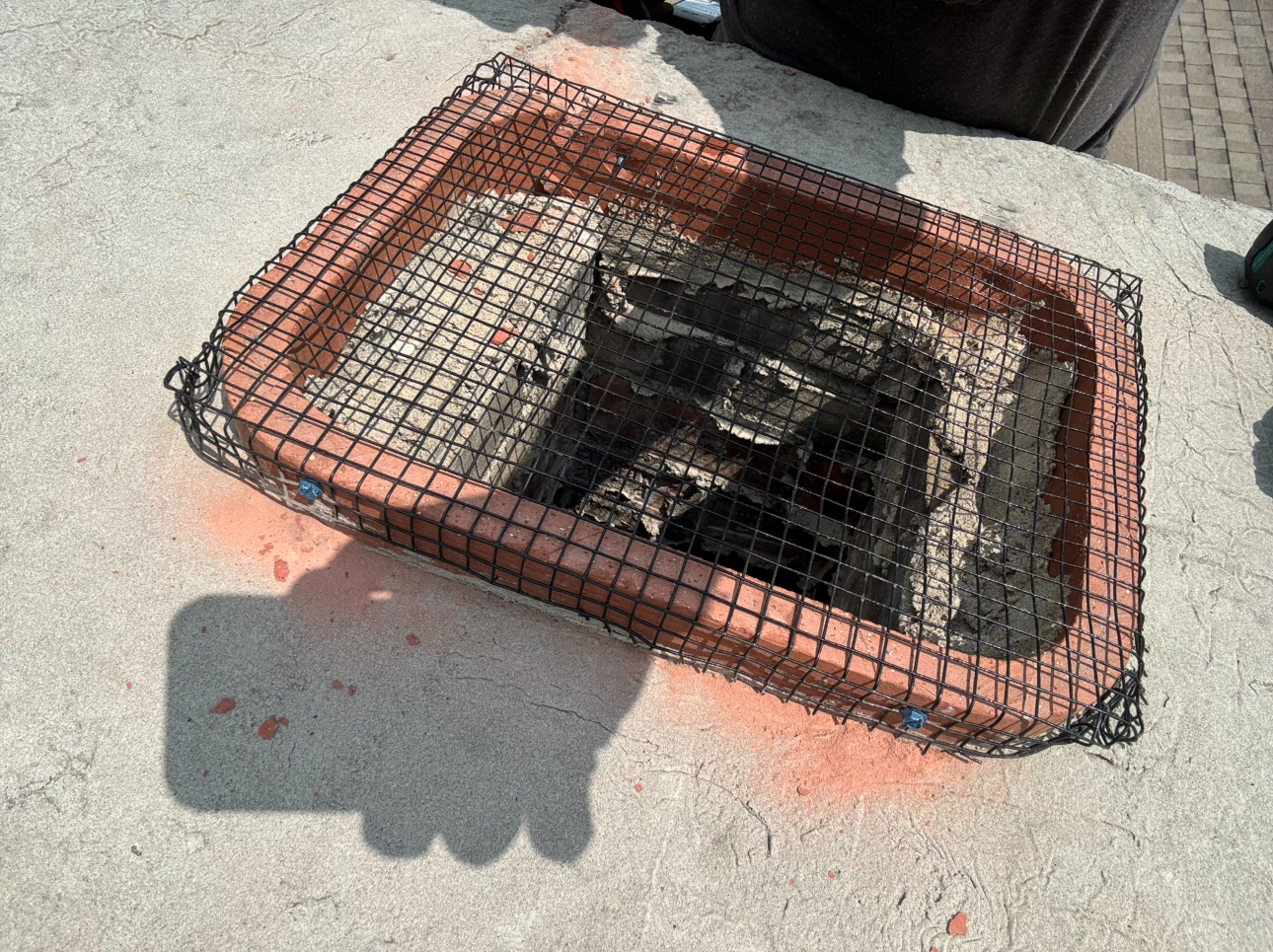 Brown rectangular planter with mesh covering, filled with dark material, on a concrete surface.