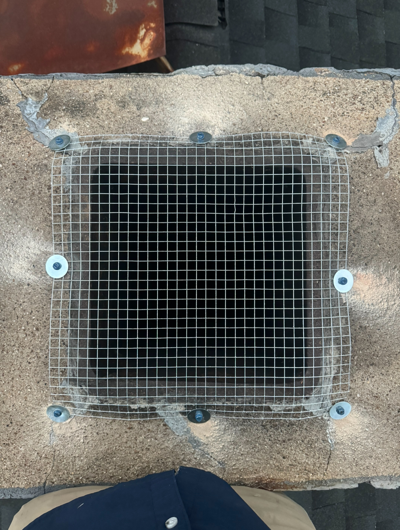 Chimney with a metal mesh covering, secured by bolts, on a roof.