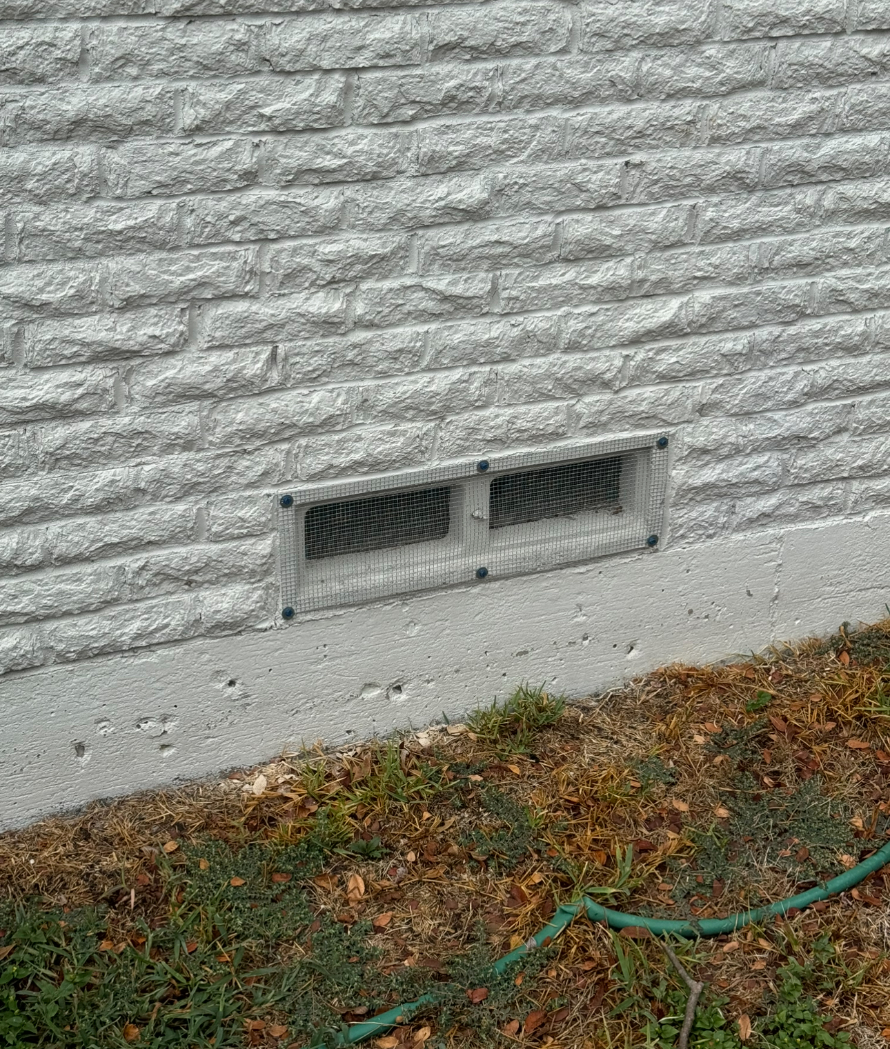 Gray stone wall with a rectangular vent covered in mesh. Grass and a hose below.