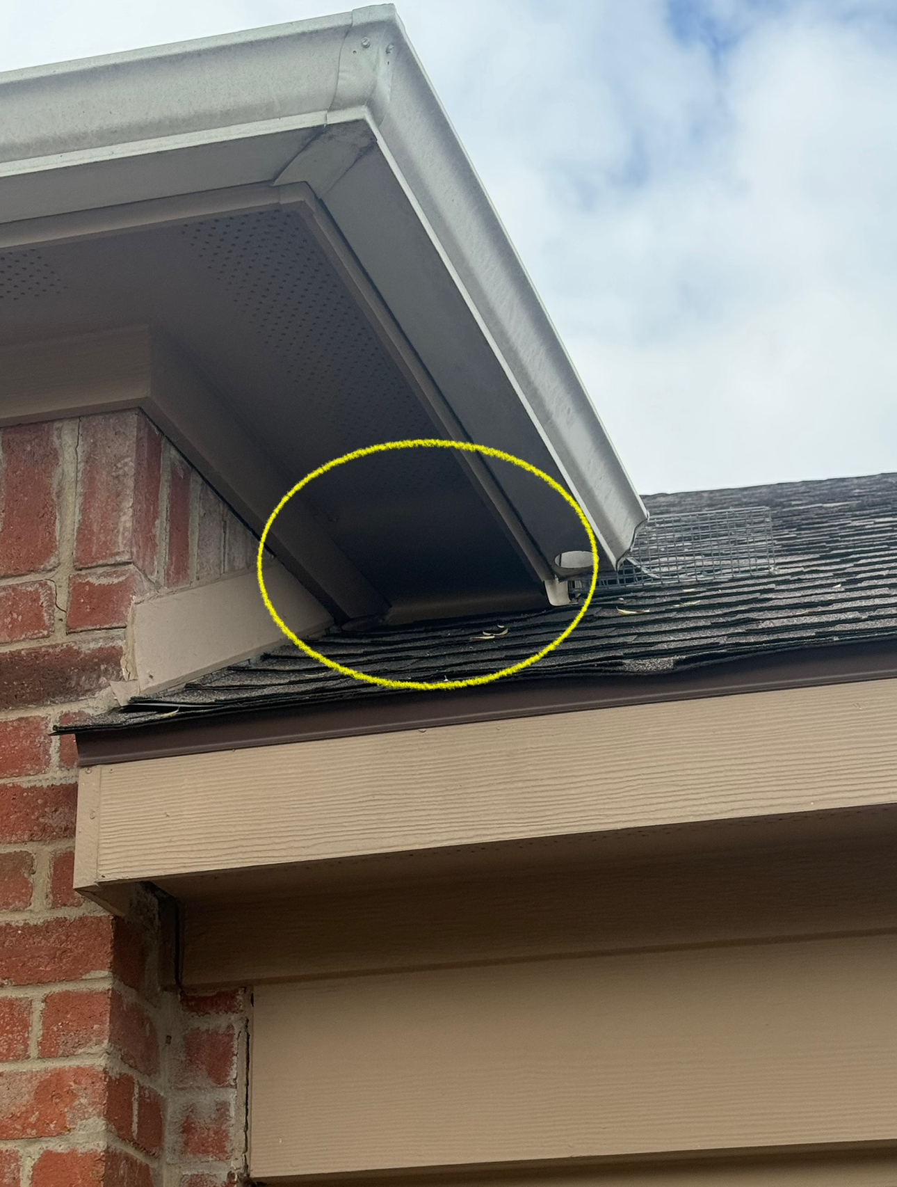 Close-up of house roof corner; yellow circle highlights possible wood damage. Brown shingles, tan trim, brick.