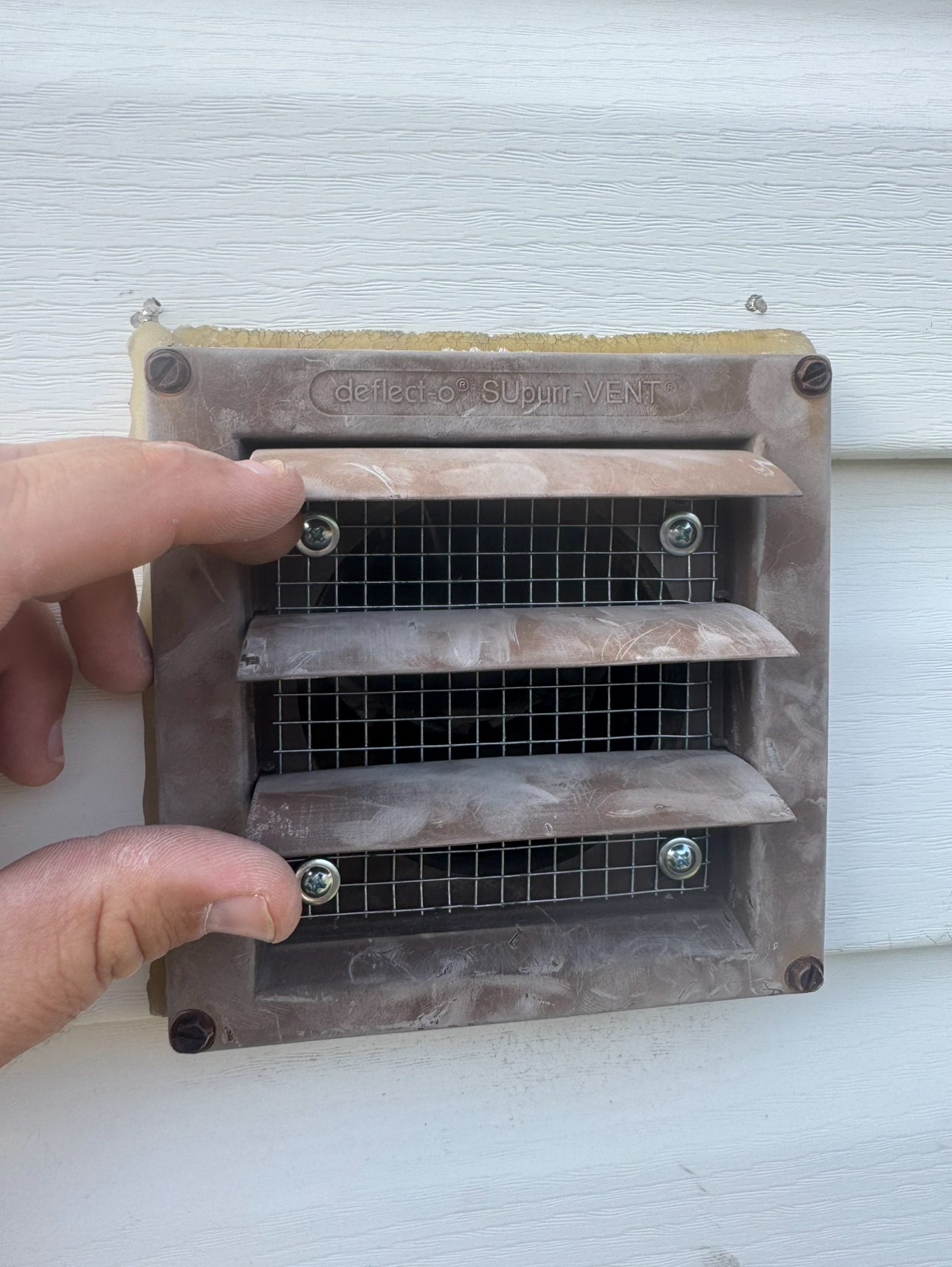Hand pointing at a brown vent on white siding. Vent has wire mesh and louvers.