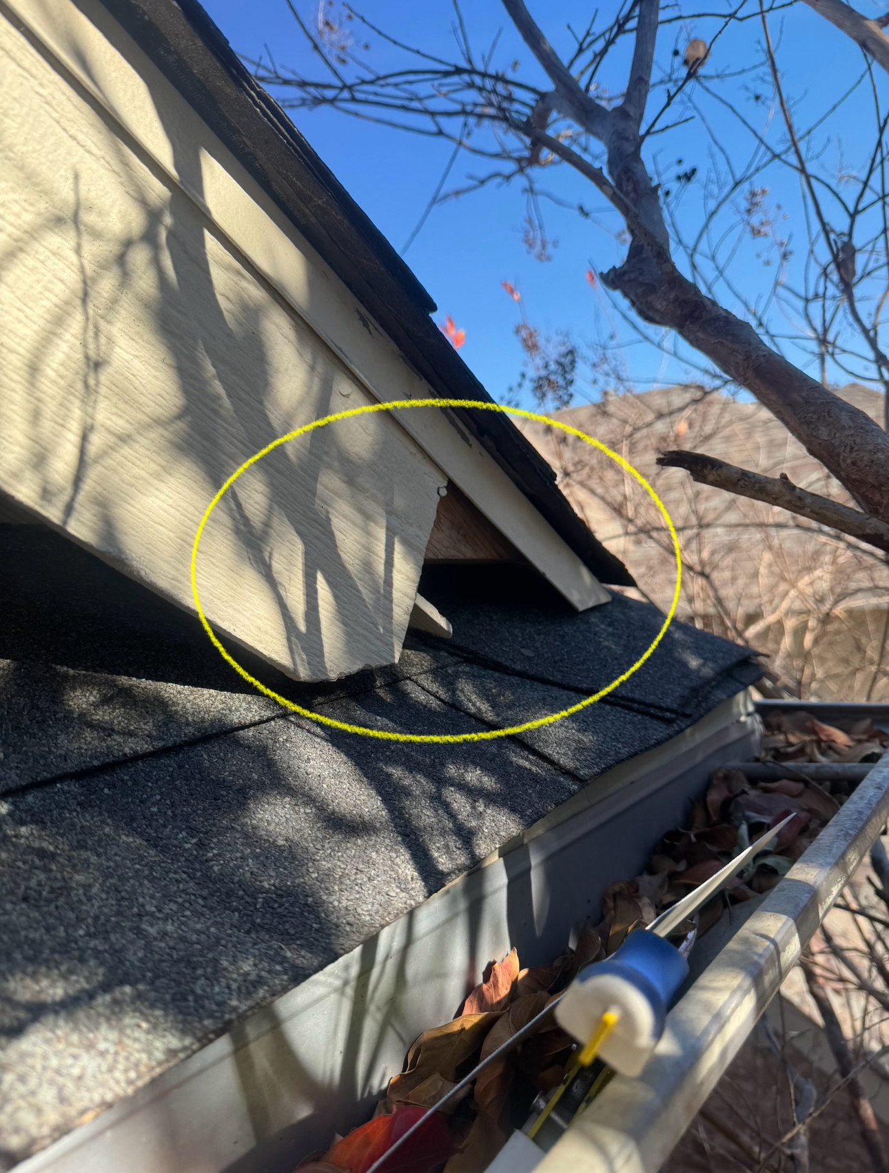 Damaged roof corner with missing siding; dark shingles, clear sky.