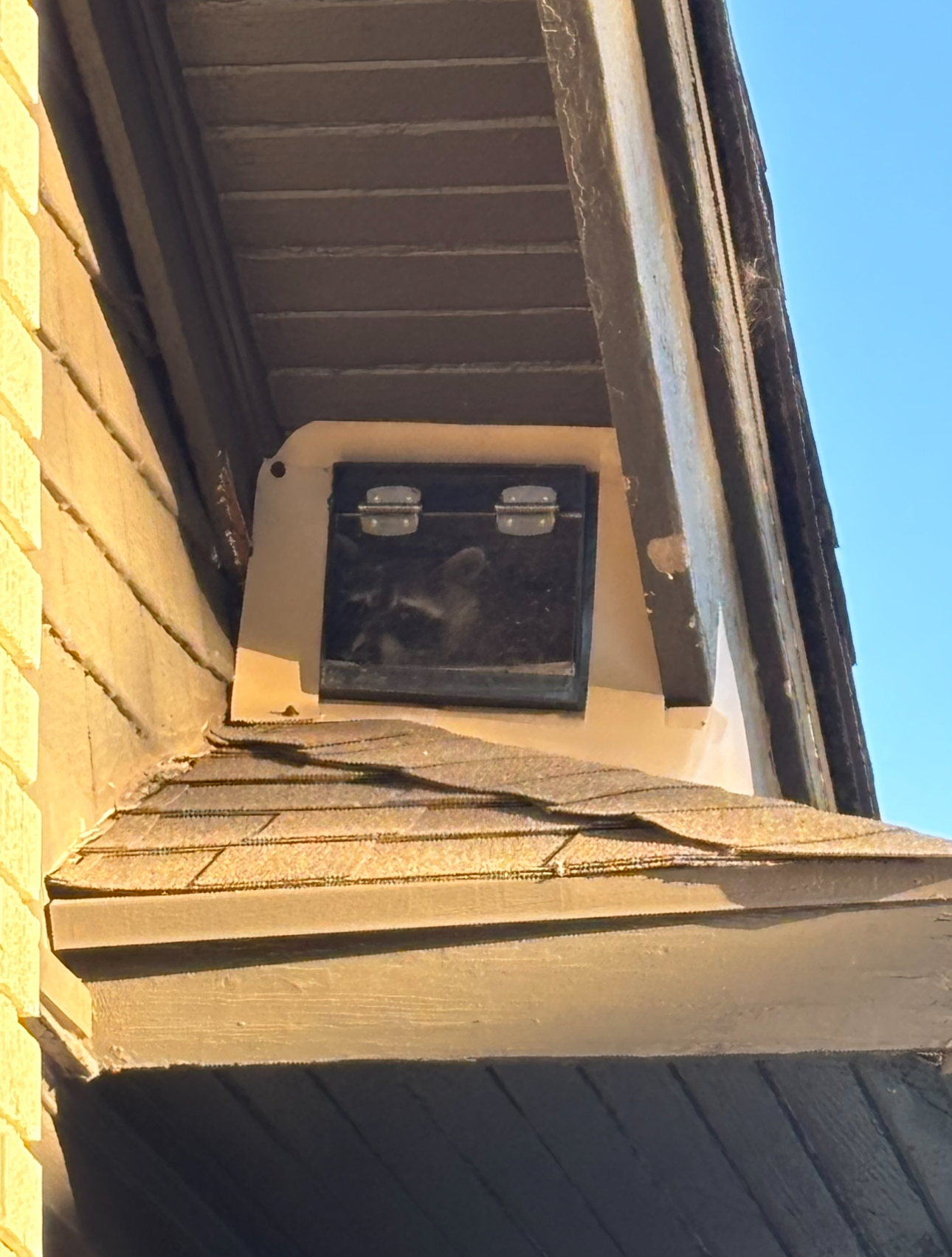 Black box with a hinged door mounted on a building's exterior, between the roof and siding.