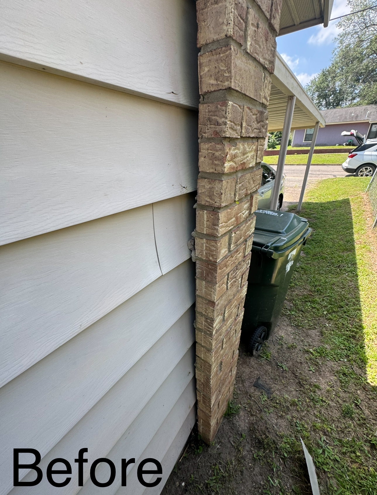 White siding next to a brown brick chimney with a crack. A green trash can sits nearby on grass.