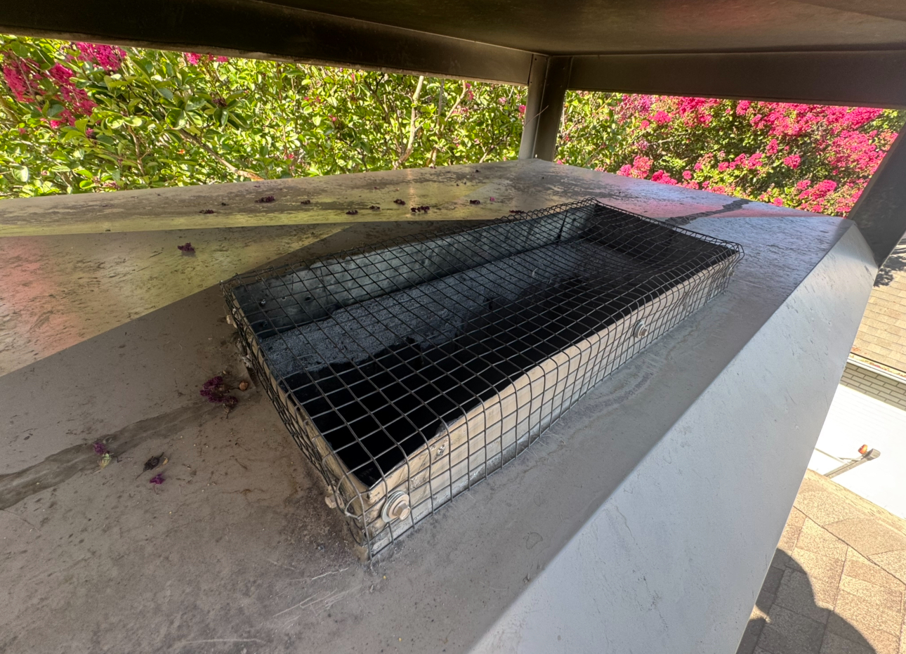 Metal mesh chimney cap on a concrete surface, viewed from above. Green foliage and pink flowers in the background.
