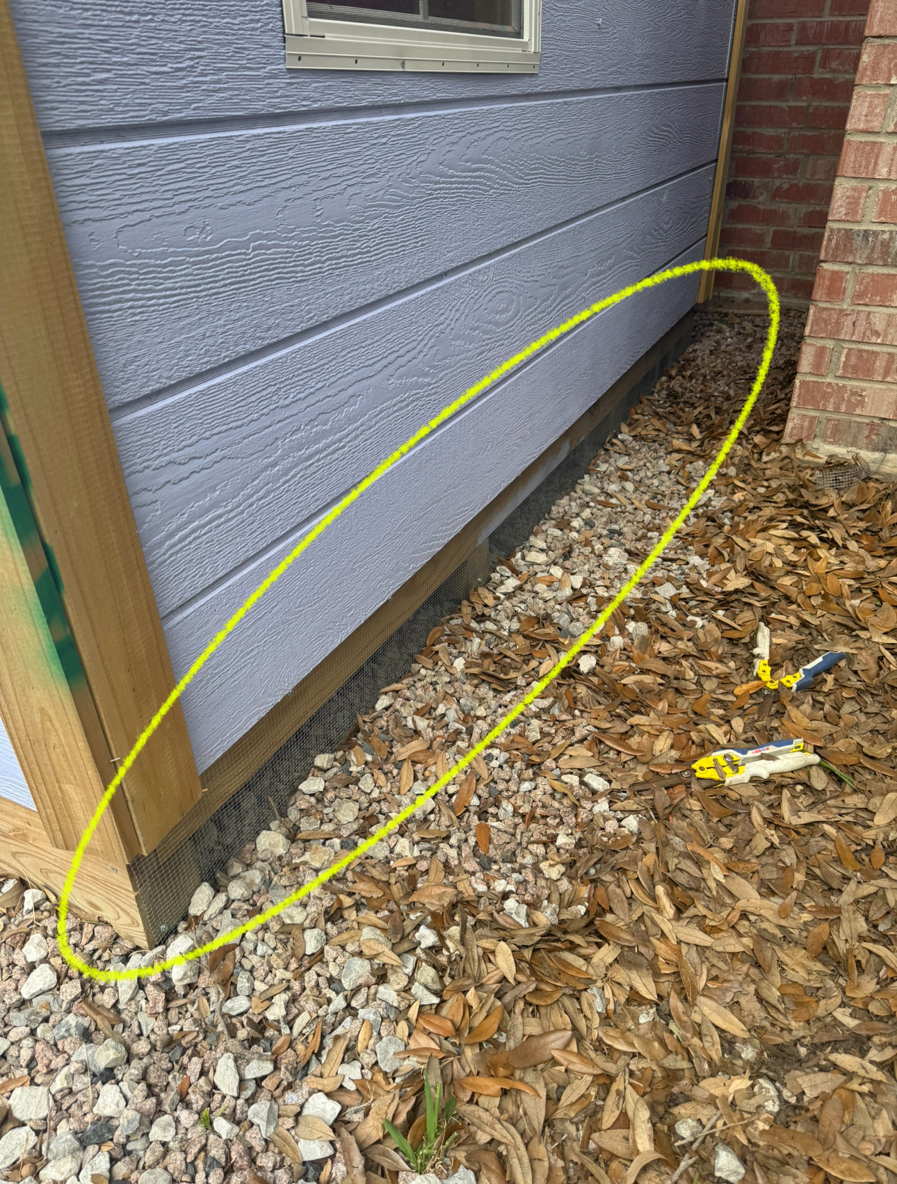 Gray siding meets gravel and leaves at the base of a building, near a brick wall.