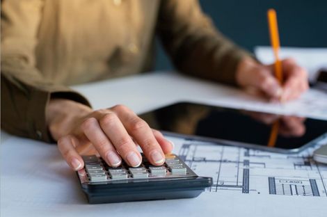 Person calculating with a calculator, writing with a pencil. A tablet, documents, and pen are visible on the white desk.