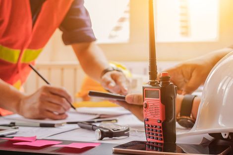 Construction workers at a table, one writing on blueprints, another holding a phone. A walkie-talkie and hard hat are also on the table.