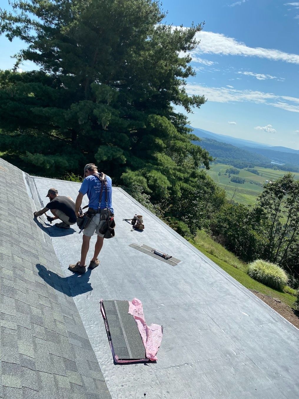 Two people working on a sloped gray roof beside trees and a wide green valley.