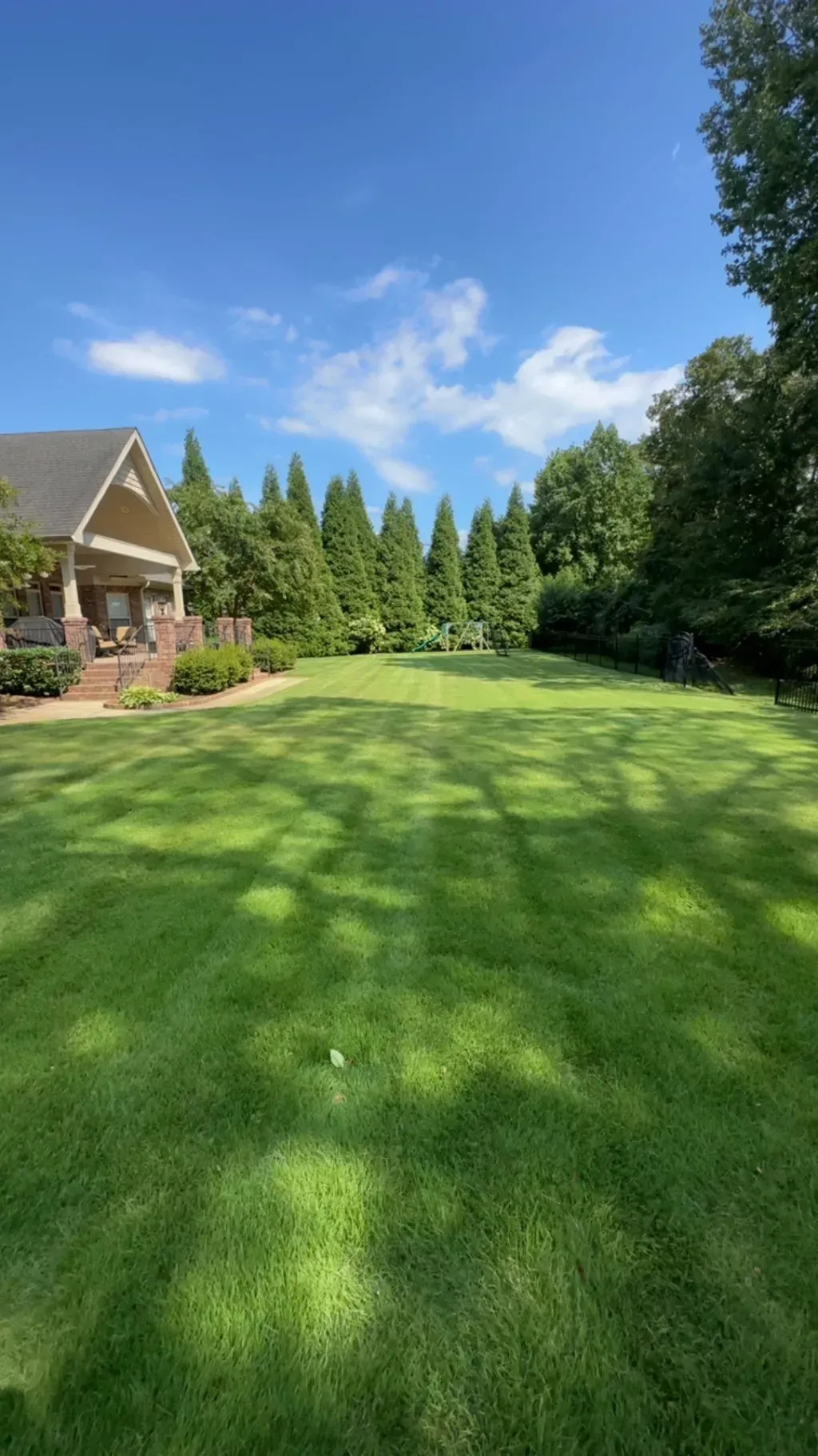 Lush green lawn with a house on the left and trees in the distance under a bright blue sky.