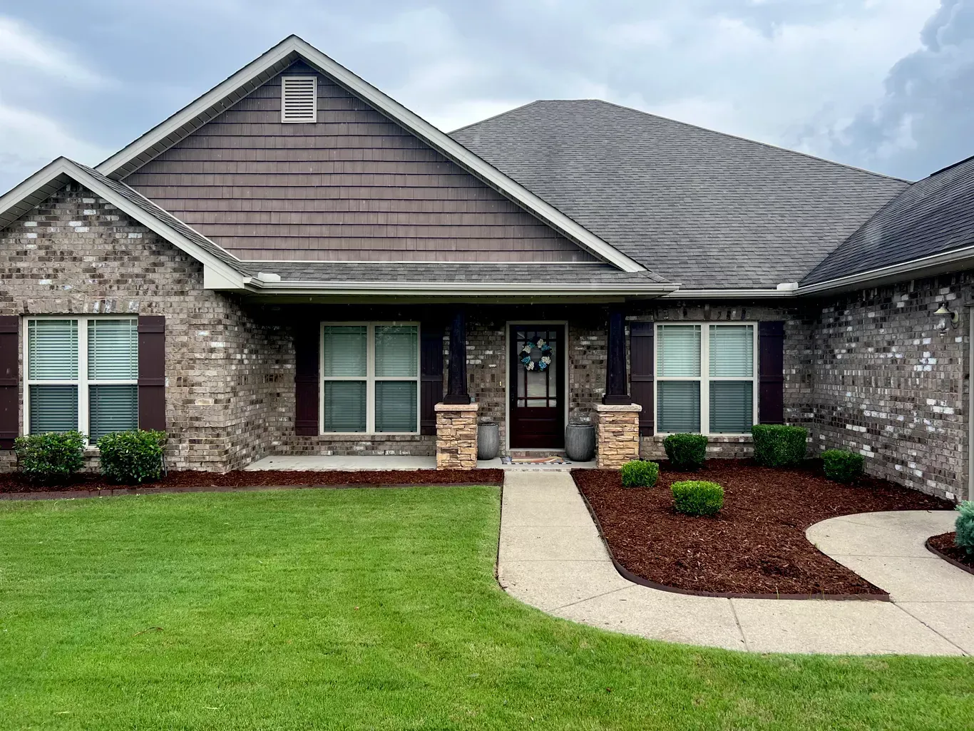 Brick house with brown roof and shutters, a concrete walkway, and brown mulch landscaping.