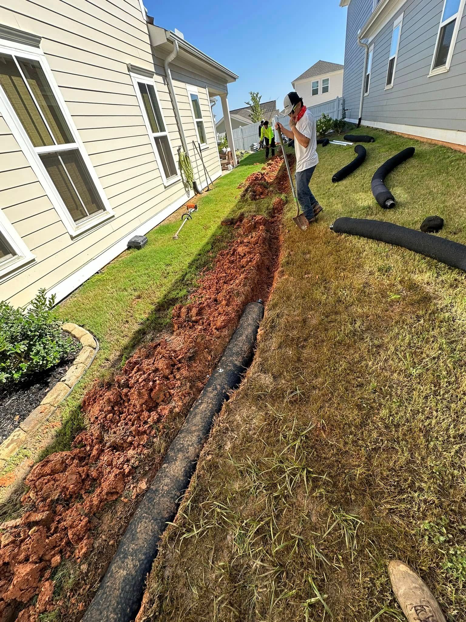 Workers installing drainage pipe in a narrow trench between houses; brown soil, green grass, blue sky.