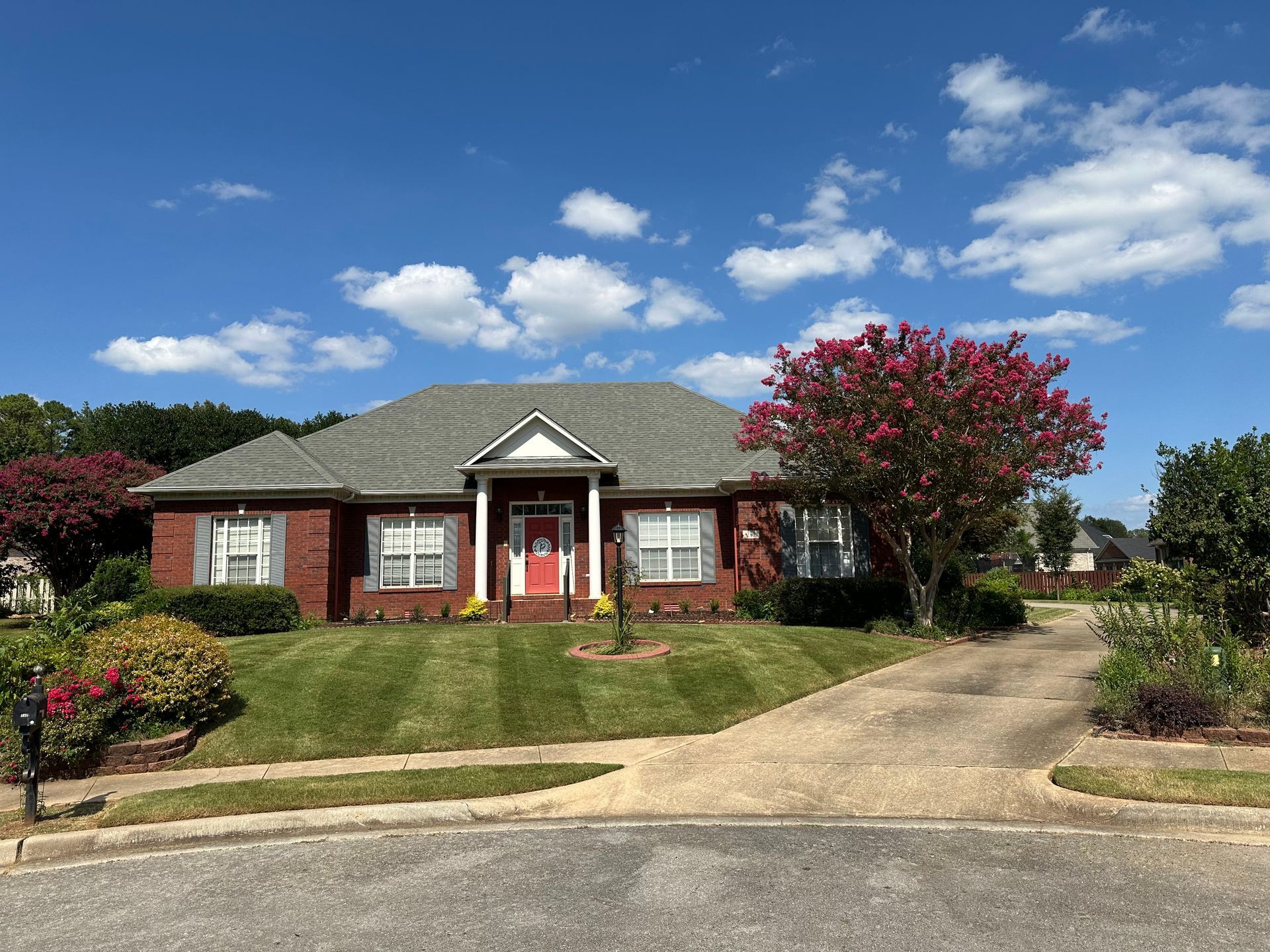 Brick house with a red door, manicured lawn, and a Crape Myrtle tree under a bright blue sky with puffy clouds.