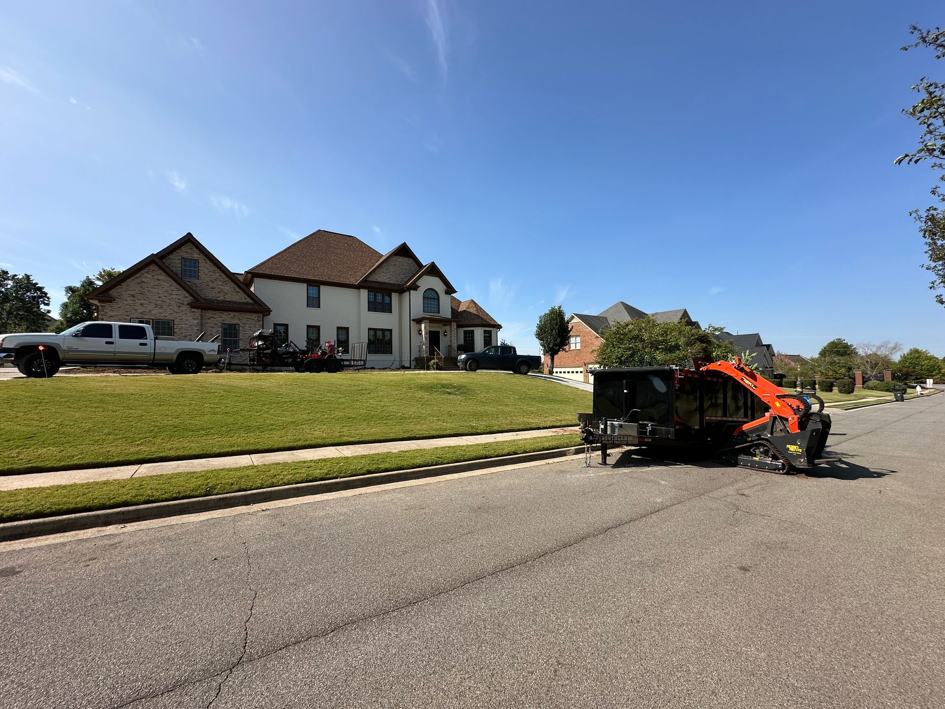 A street scene with a large house, truck, trailer, and construction equipment on a sunny day.
