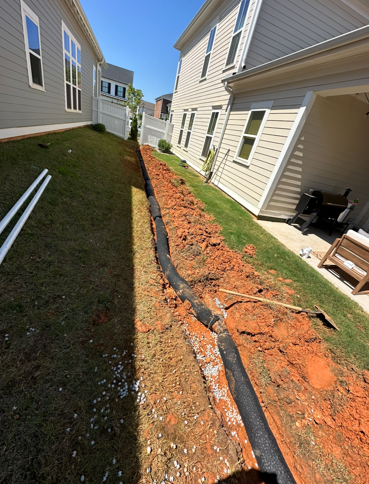 Ditch with black drainage pipe running between houses on a grassy hill. Red clay soil exposed.