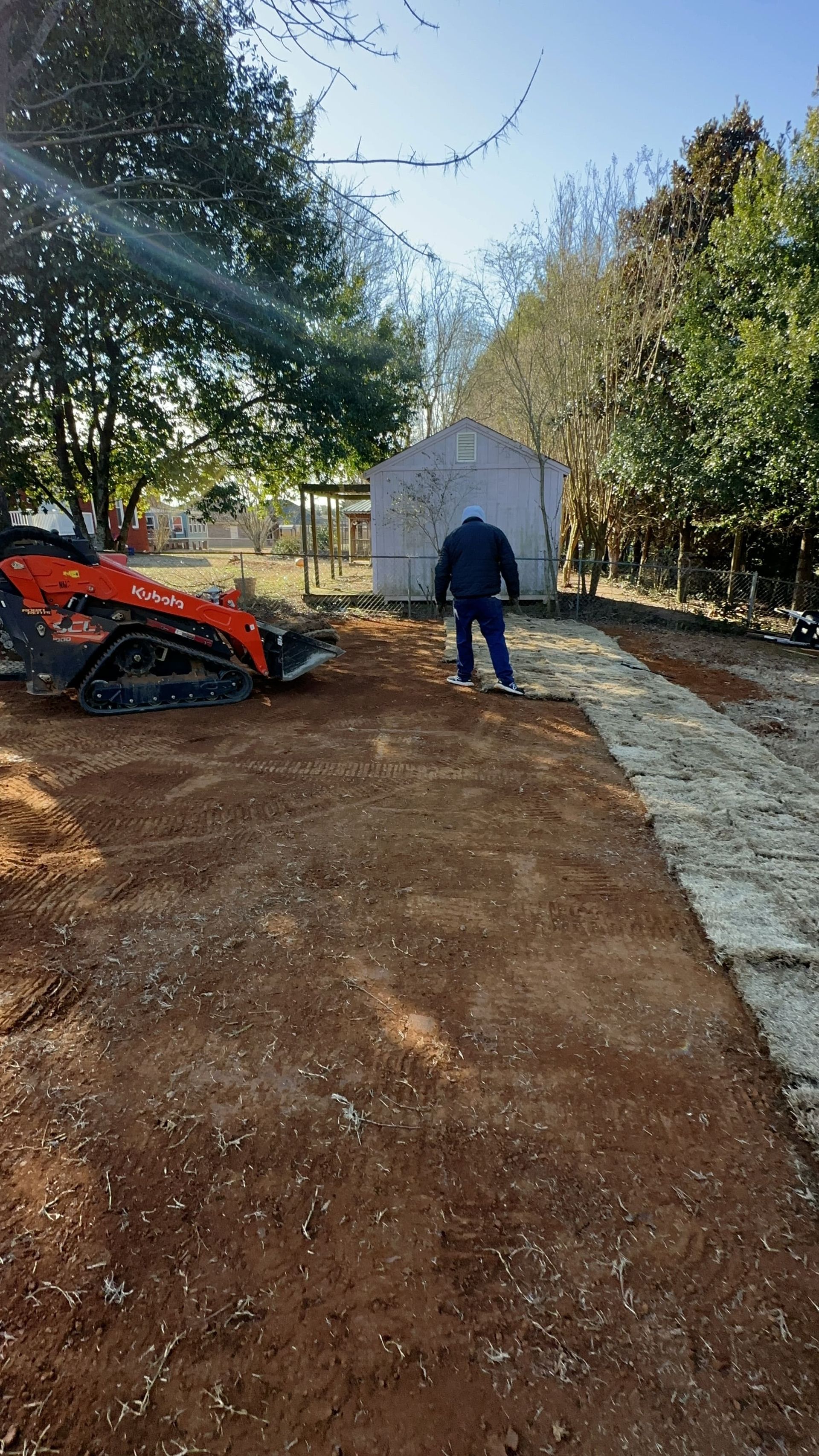 Person standing on dirt path near an orange skid steer and a white shed on a sunny day.