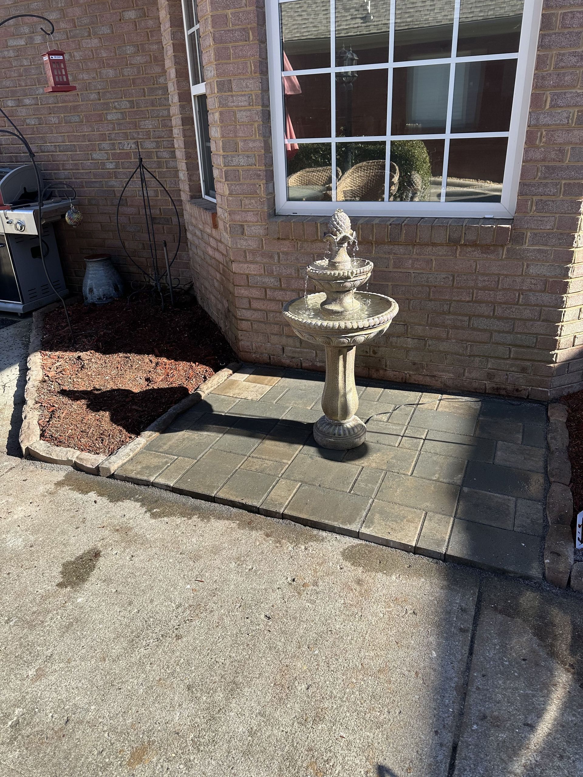 A stone fountain on a paved area in front of a brick building, next to a small garden bed.