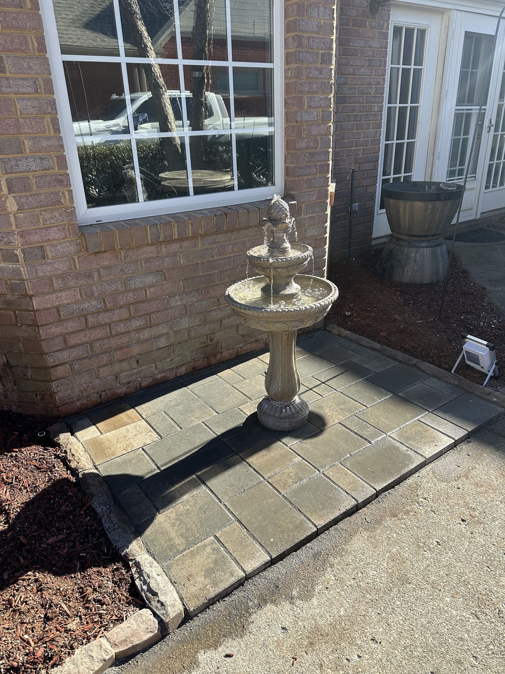 Fountain on a brick patio beside a building. Brown brick wall, white framed window, and gravel walkway.