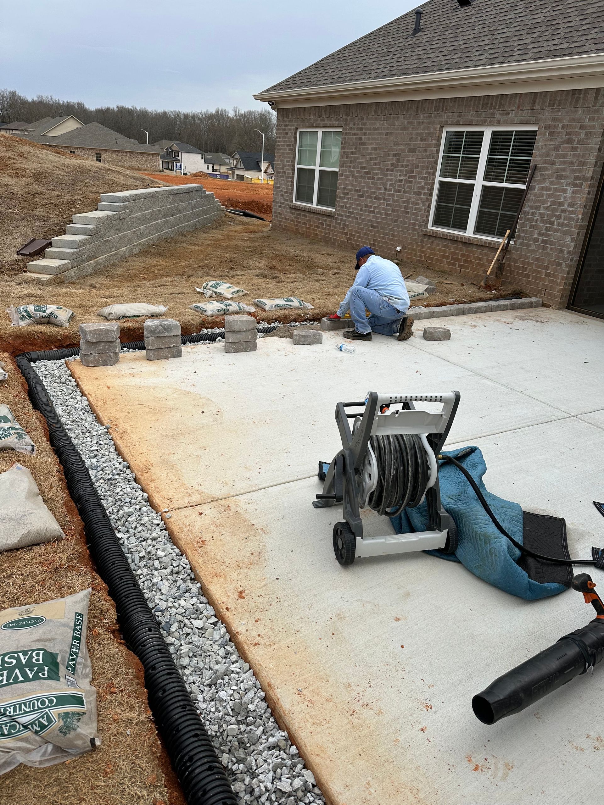 Construction worker laying pavers near a patio with a trench drain; a brick house in the background.