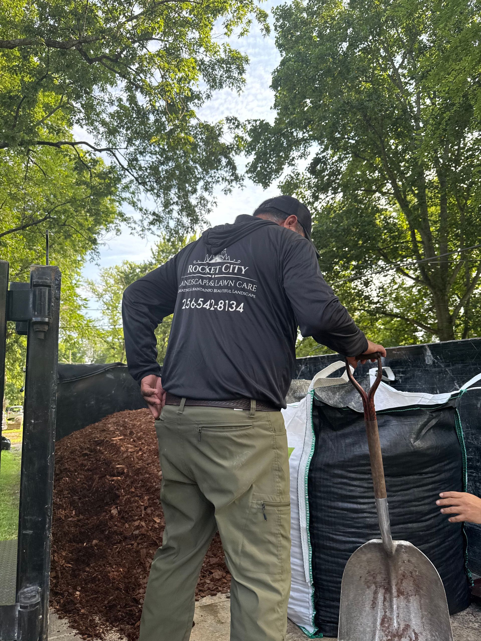 Man shoveling mulch from a truck bed. Wearing a black sweatshirt, green pants. Bags of mulch and trees in the background.