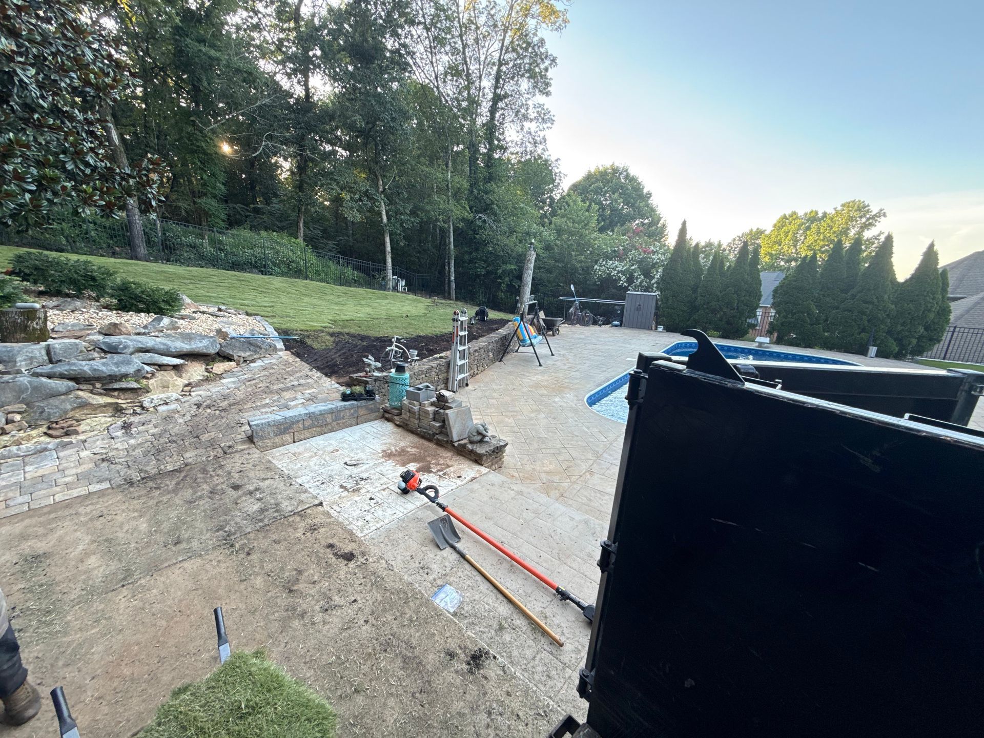Construction site with workers, dirt, stone walls, and a pool; sunny outdoors.
