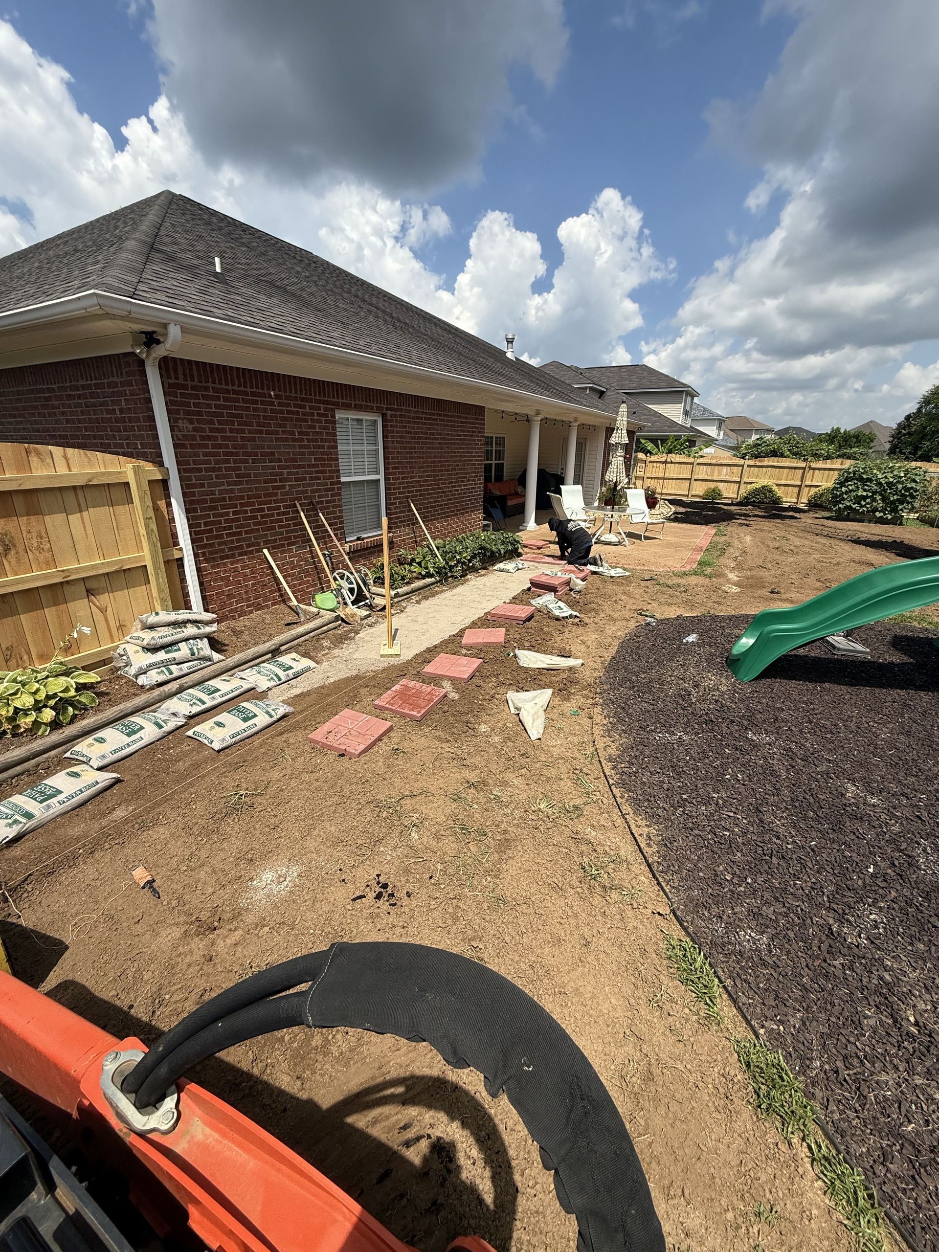 Backyard landscaping with brick house, mulch, red stepping stones, and green playset under cloudy skies.