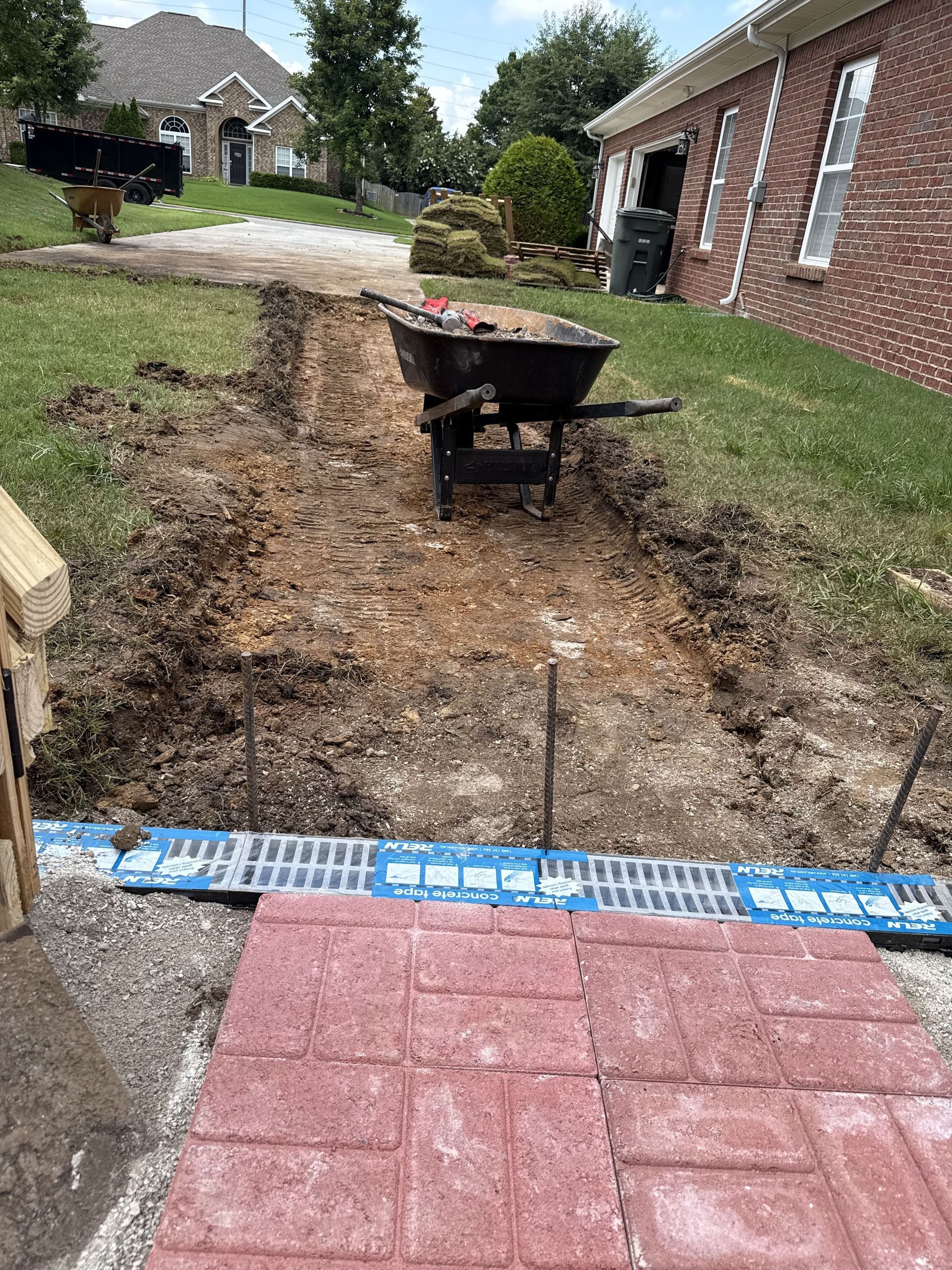 Pathway construction with a wheelbarrow, red bricks, and a drainage system.