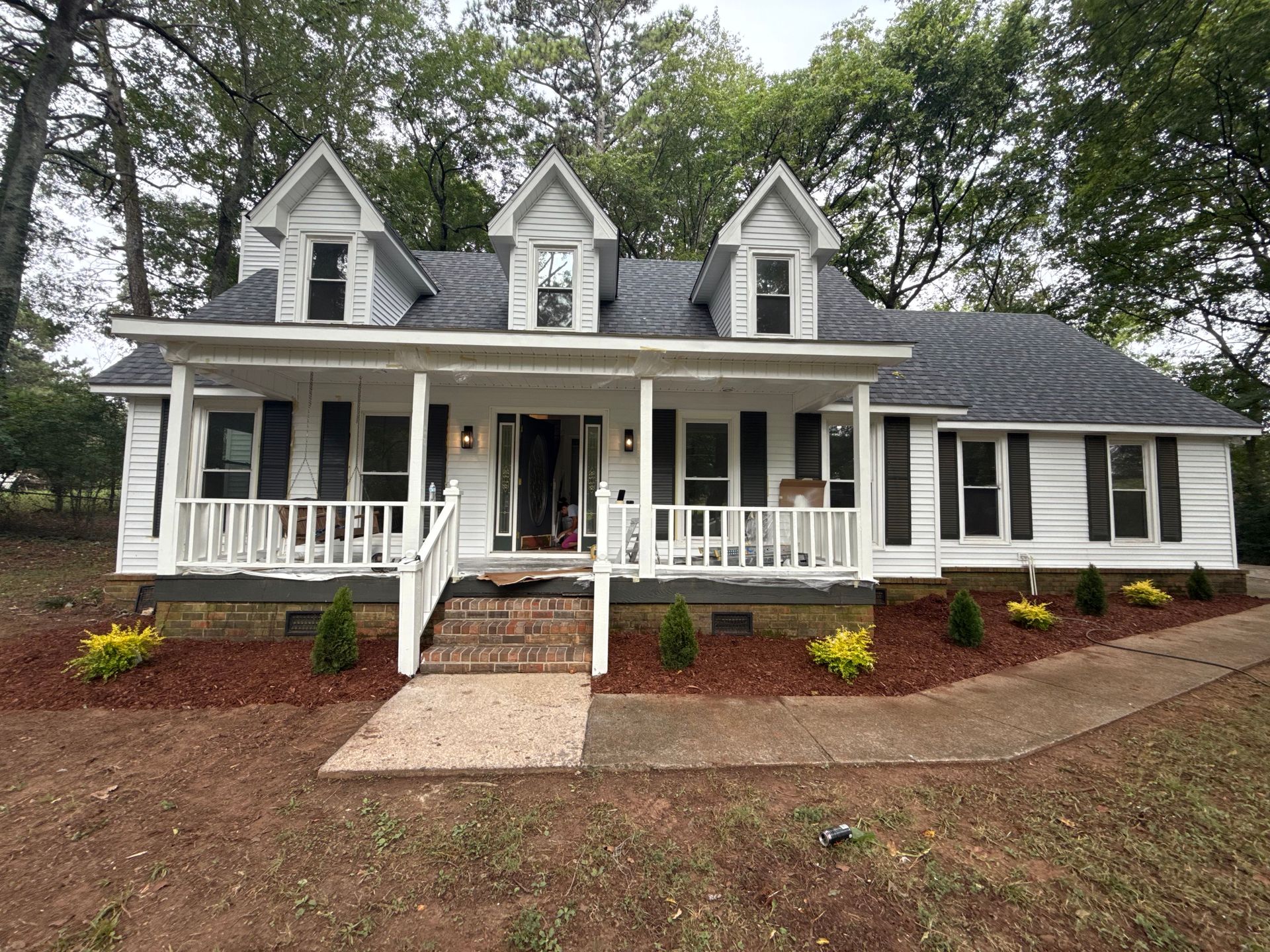 White house with porch, black shutters, and three dormers, set in a yard with landscaping.