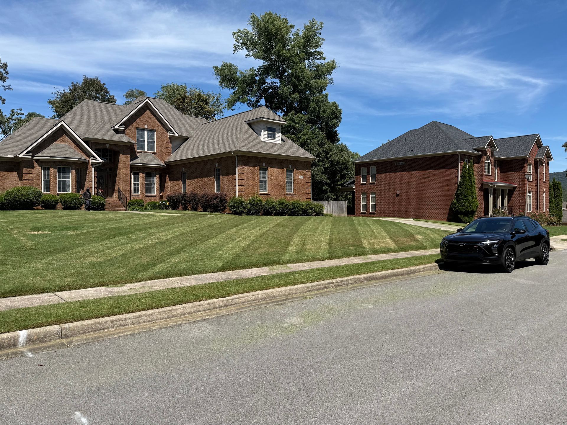 Two brick houses with well-manicured lawns and a black car parked on a street on a sunny day.