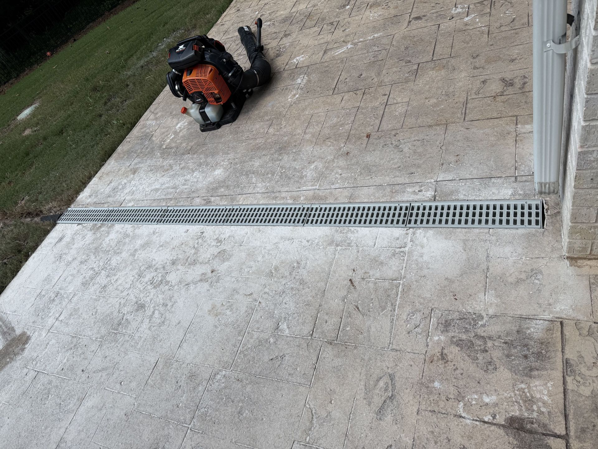 A concrete patio with a long drain, a backpack, and a grassy hill on the left.