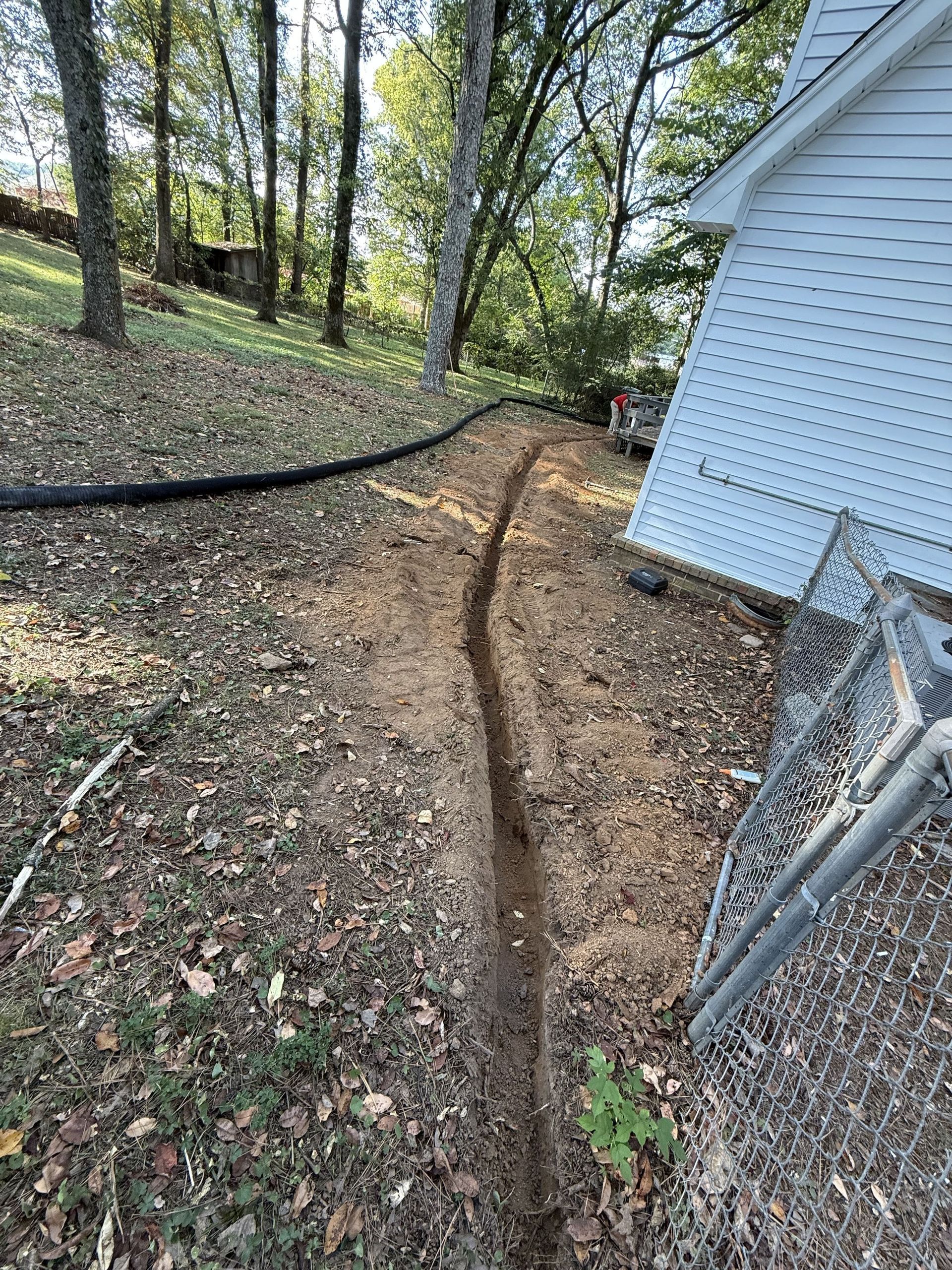Trench dug in yard near a house, for a black pipe. Dirt, leaves, trees in background.