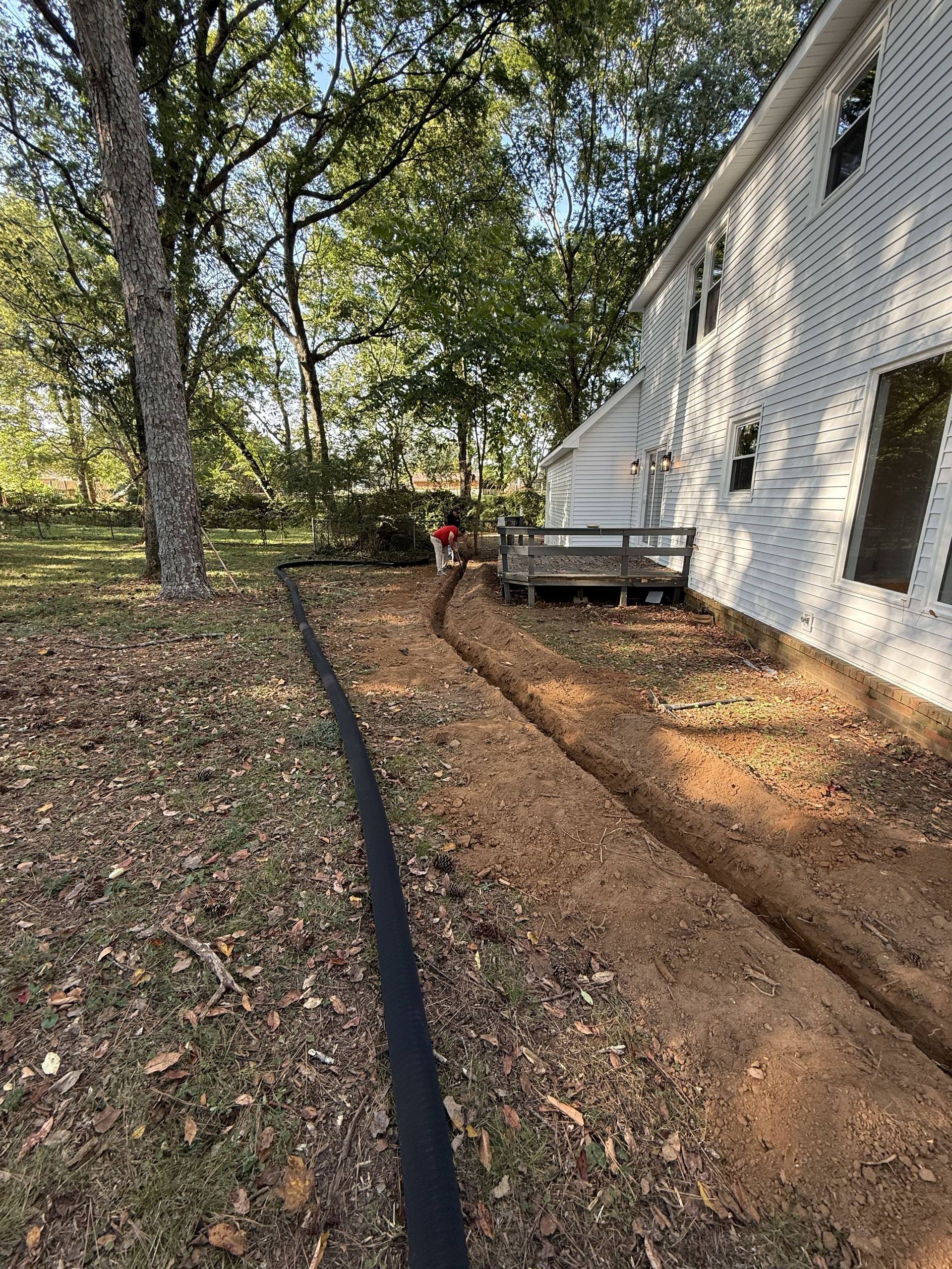 A trench dug in a yard, with a house on the right and trees in the background.