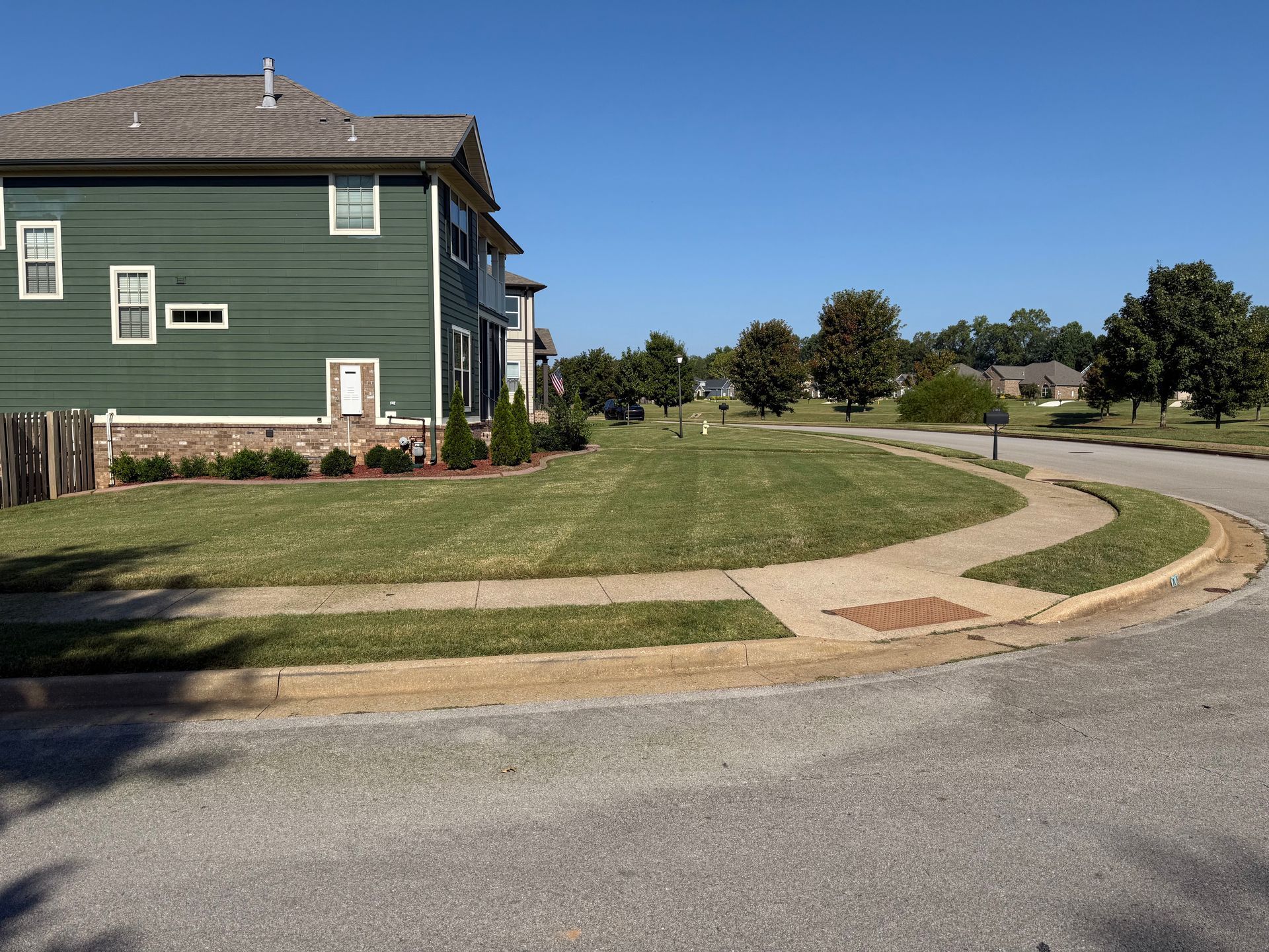 Green house on a corner lot with freshly mowed lawn, sidewalk, and street under a blue sky.