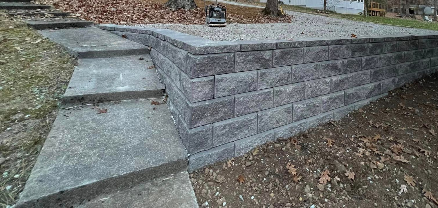 Concrete steps lead to a retaining wall built of gray blocks, surrounded by dirt and leaves.