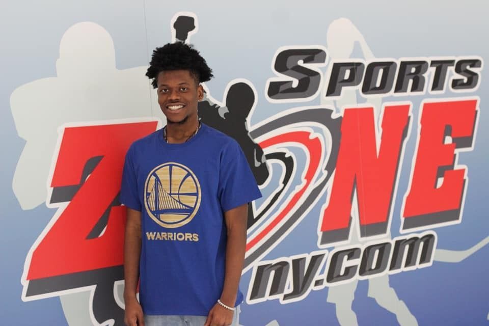A young man in a blue Warriors shirt smiles in front of a Sports Zone backdrop.