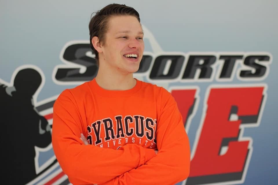 Man in orange Syracuse sweatshirt smiles, arms crossed, in front of 