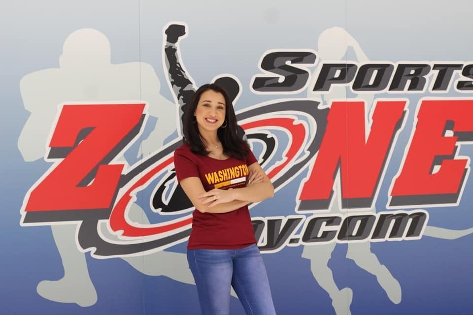 Woman with crossed arms, smiling, wearing maroon shirt and jeans in front of a Sports Zone sign.