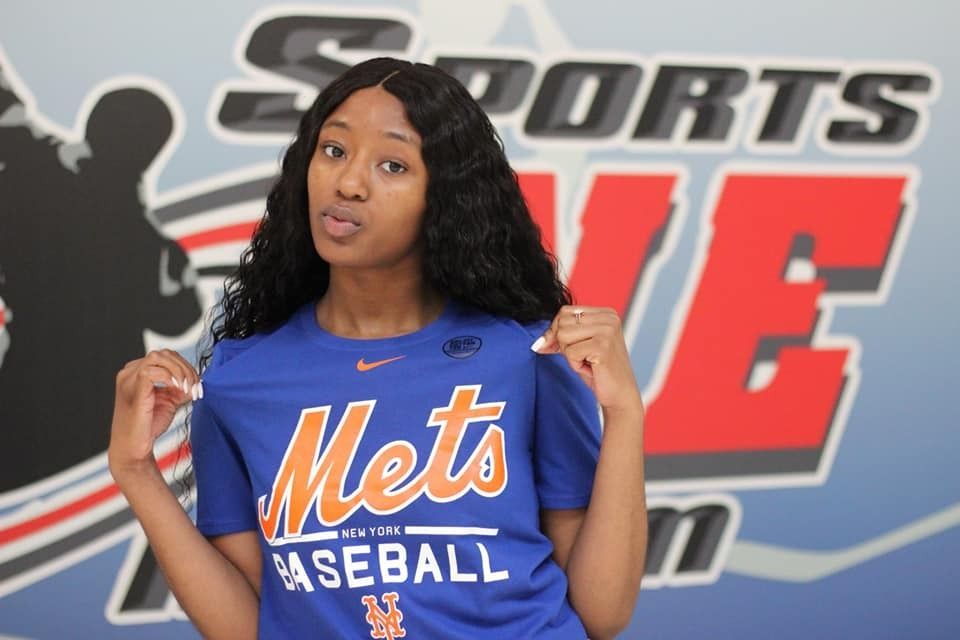 Woman in a blue Mets shirt, holding it open, in front of a sports logo backdrop.